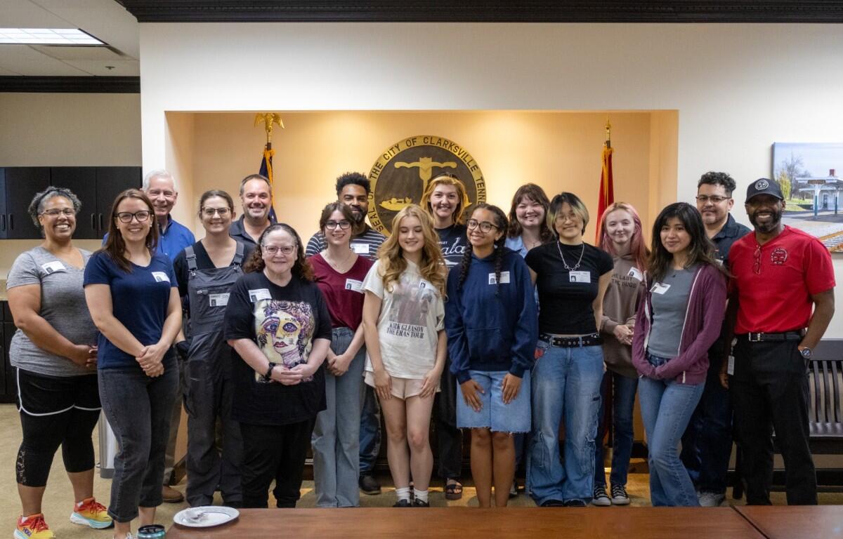Group of about 20 diverse adults and teens posing for a photo in a city government room, with seals and flags in the background, smiling at the camera.