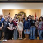 Group of about 20 diverse adults and teens posing for a photo in a city government room, with seals and flags in the background, smiling at the camera.