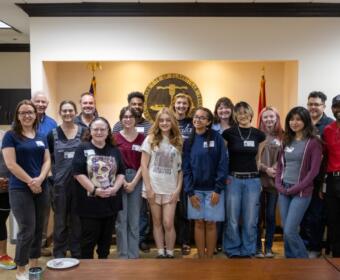 Group of about 20 diverse adults and teens posing for a photo in a city government room, with seals and flags in the background, smiling at the camera.