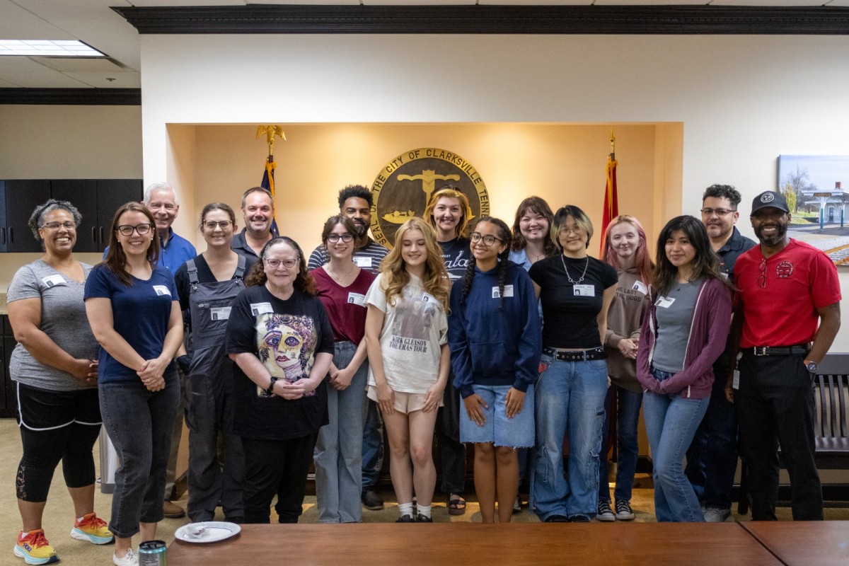 Group of about 20 diverse adults and teens posing for a photo in a city government room, with seals and flags in the background, smiling at the camera.