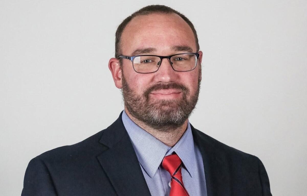 Portrait of a man with short hair and beard, wearing glasses, a navy blazer, light blue shirt, and red striped tie, looking at the camera.