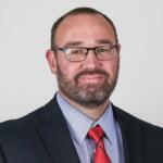 Portrait of a man with short hair and beard, wearing glasses, a navy blazer, light blue shirt, and red striped tie, looking at the camera.