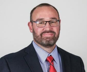 Portrait of a man with short hair and beard, wearing glasses, a navy blazer, light blue shirt, and red striped tie, looking at the camera.