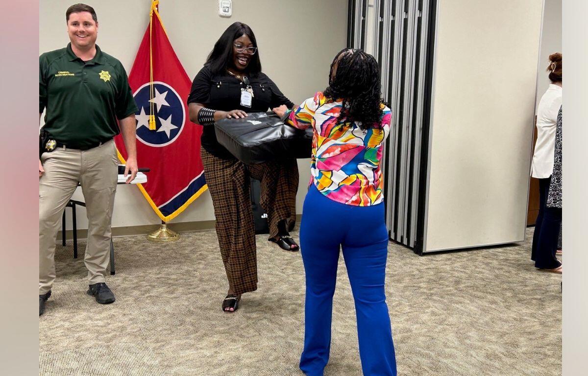 Two women exchange a black package in an office setting; a man in uniform stands nearby with a Tennessee flag in the background.