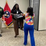 Two women exchange a black package in an office setting; a man in uniform stands nearby with a Tennessee flag in the background.