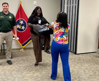 Two women exchange a black package in an office setting; a man in uniform stands nearby with a Tennessee flag in the background.