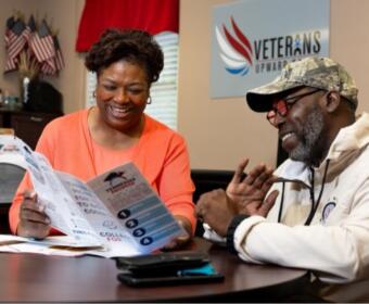 Two adults sit at a desk reading a brochure; a Veterans Upward Bound sign is on the wall behind them.