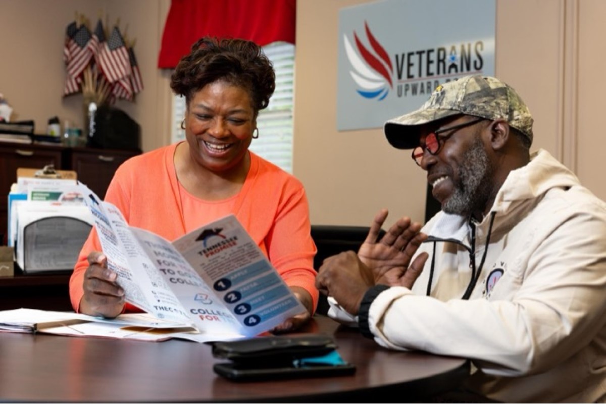 Two adults sit at a desk reading a brochure; a Veterans Upward Bound sign is on the wall behind them.