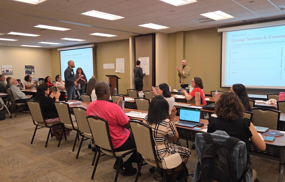 Group of adults in a workshop setting, some seated at tables applauding while a presenter stands at front with a projector screen reading 'Group Norms & Commitments'.