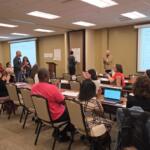 Group of adults in a workshop setting, some seated at tables applauding while a presenter stands at front with a projector screen reading 'Group Norms & Commitments'.