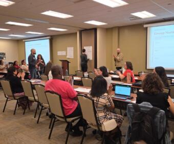 Group of adults in a workshop setting, some seated at tables applauding while a presenter stands at front with a projector screen reading 'Group Norms & Commitments'.
