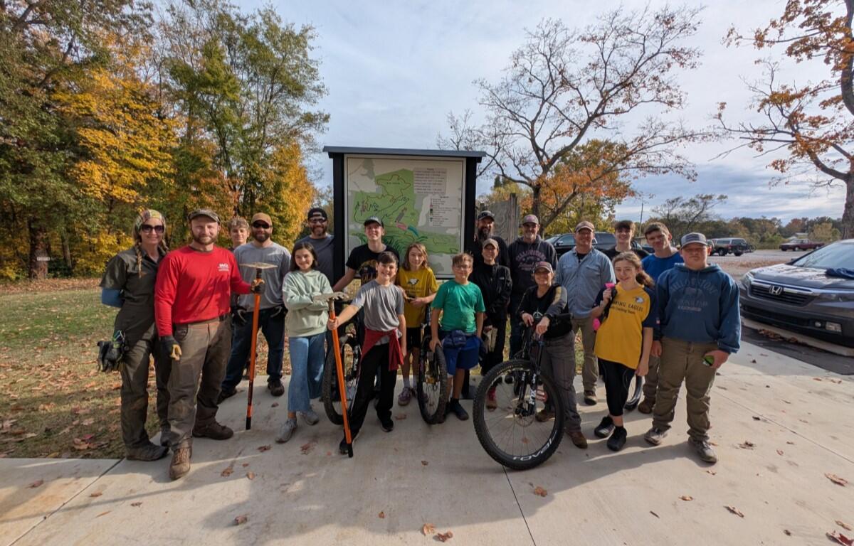 Group of people posing in front of a park map sign on a fall day, some with bikes and tools, smiling for the photo.