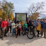 Group of people posing in front of a park map sign on a fall day, some with bikes and tools, smiling for the photo.