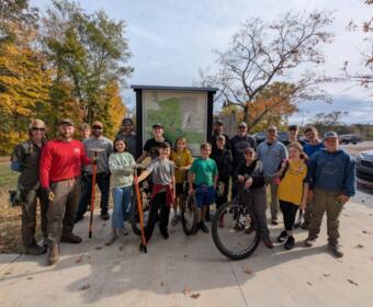 Group of people posing in front of a park map sign on a fall day, some with bikes and tools, smiling for the photo.