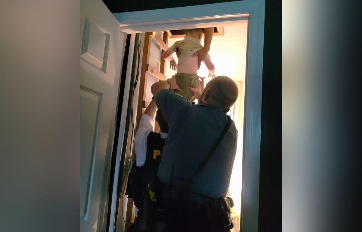 Two police officers lift a child up through an open doorway into a brightly lit room in a stairwell area.