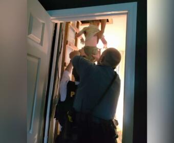 Two police officers lift a child up through an open doorway into a brightly lit room in a stairwell area.