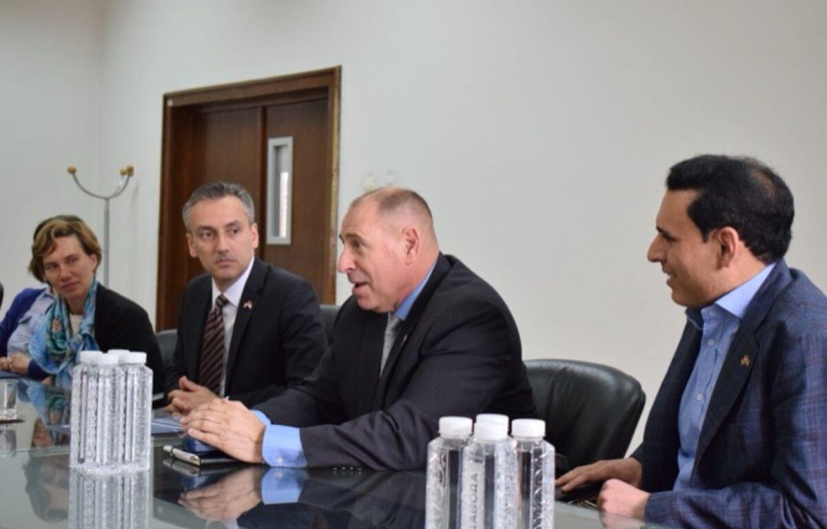 Four professionals seated at a glass conference table during a formal meeting, with water bottles visible in the foreground.