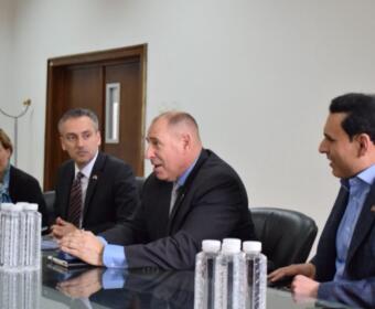 Four professionals seated at a glass conference table during a formal meeting, with water bottles visible in the foreground.