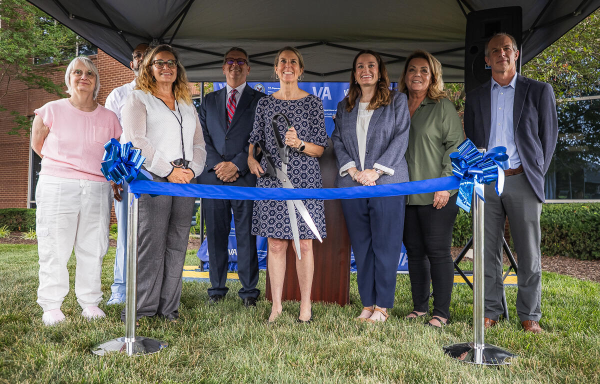 Group of eight people standing under a tent, smiling, about to cut a blue ribbon at a ceremonial ribbon-cutting event outdoors.