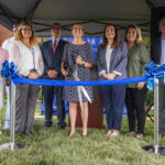 Group of eight people standing under a tent, smiling, about to cut a blue ribbon at a ceremonial ribbon-cutting event outdoors.