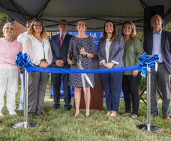 Group of eight people standing under a tent, smiling, about to cut a blue ribbon at a ceremonial ribbon-cutting event outdoors.