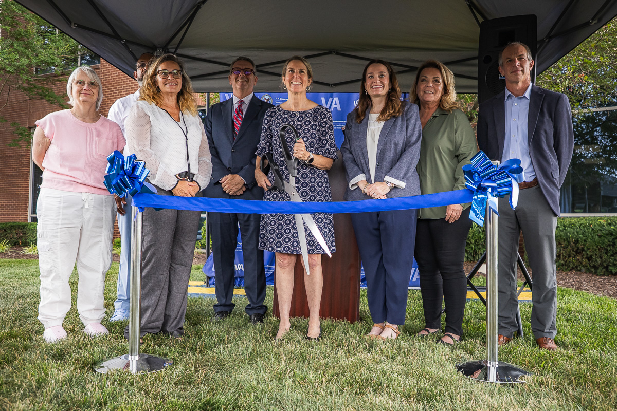 Group of eight people standing under a tent, smiling, about to cut a blue ribbon at a ceremonial ribbon-cutting event outdoors.