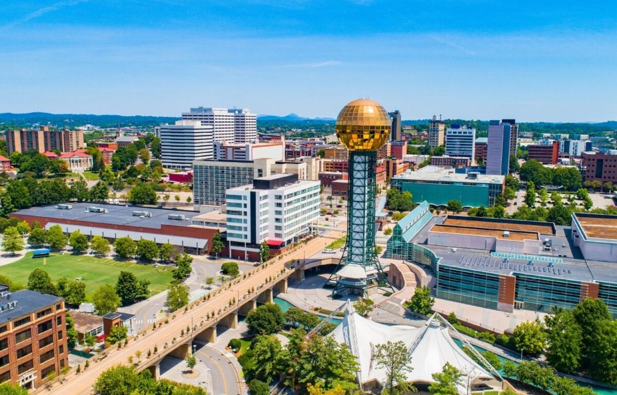 Aerial view of a city with a tall tower topped by a gold reflective sphere, near a white tent structure and modern buildings.