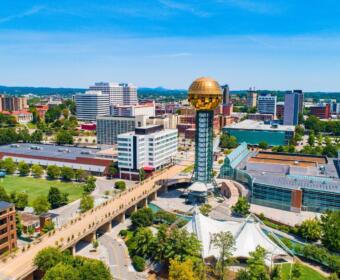 Aerial view of a city with a tall tower topped by a gold reflective sphere, near a white tent structure and modern buildings.