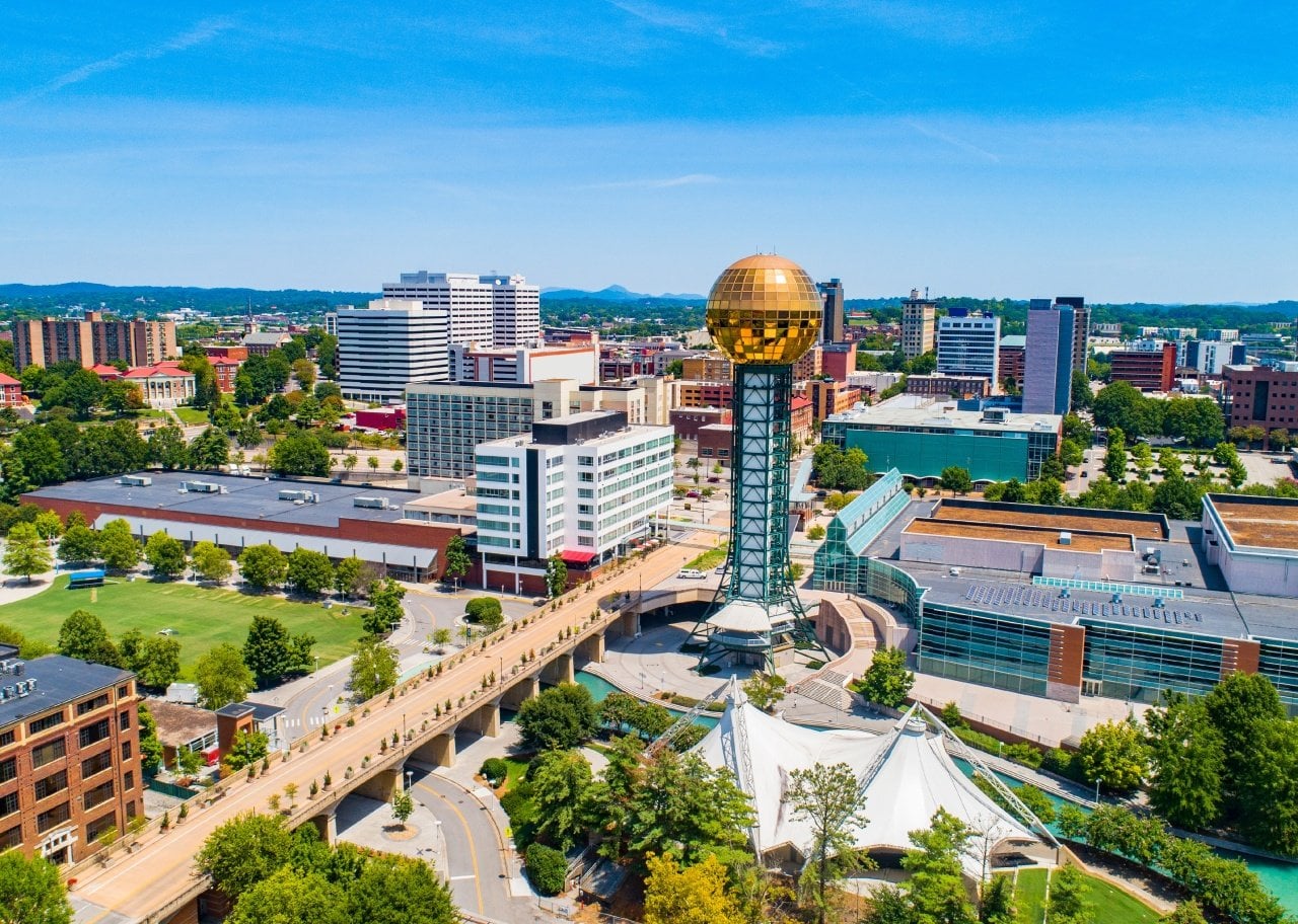 Aerial view of a city with a tall tower topped by a gold reflective sphere, near a white tent structure and modern buildings.