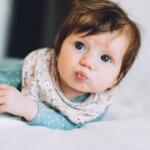 Baby lying on a bed, looking at the camera and puckering lips.