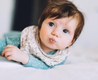 Baby lying on a bed, looking at the camera and puckering lips.