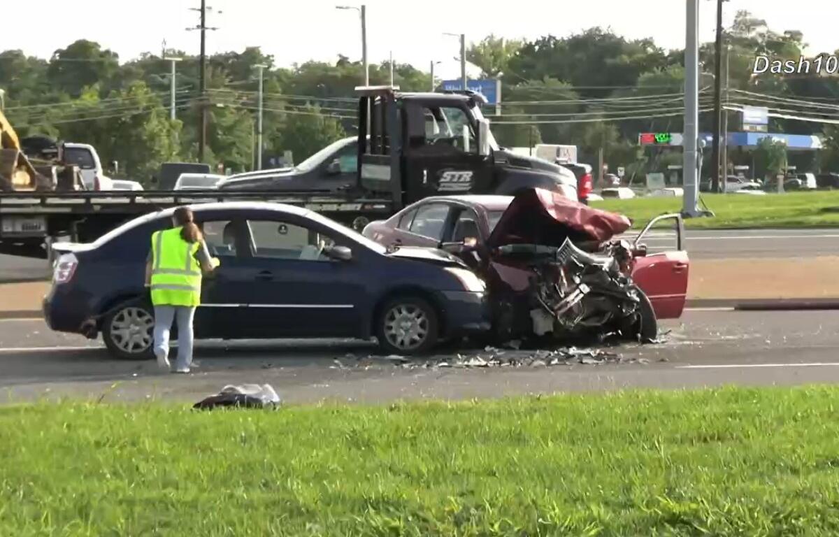 Two-car crash on a highway: a blue sedan rear‑ended by a red car with extensive front damage; a worker in a neon vest stands nearby on the road.