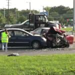 Two-car crash on a highway: a blue sedan rear‑ended by a red car with extensive front damage; a worker in a neon vest stands nearby on the road.