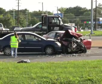Two-car crash on a highway: a blue sedan rear‑ended by a red car with extensive front damage; a worker in a neon vest stands nearby on the road.