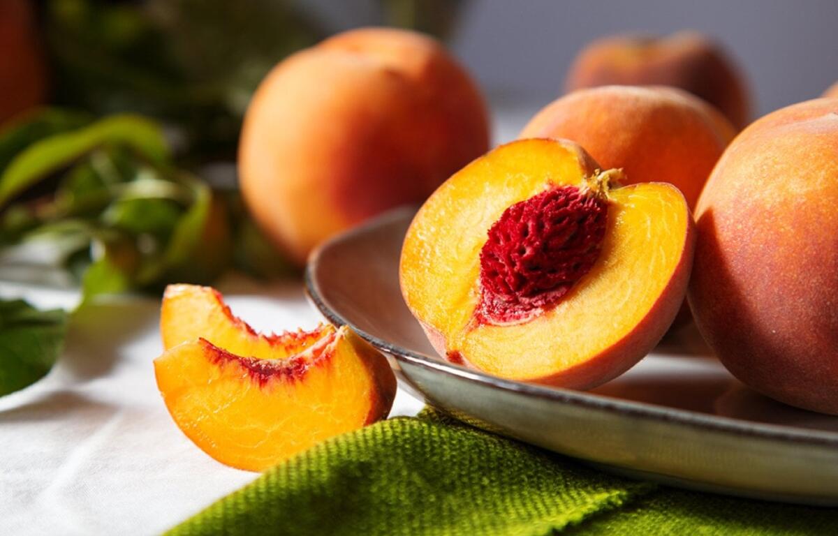 Halved ripe peach on a metal plate, bright orange flesh around a red seed, with whole peaches in the background.