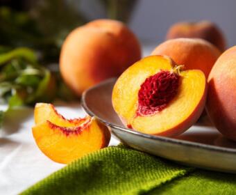 Halved ripe peach on a metal plate, bright orange flesh around a red seed, with whole peaches in the background.
