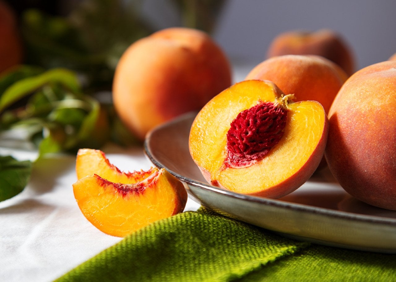 Halved ripe peach on a metal plate, bright orange flesh around a red seed, with whole peaches in the background.