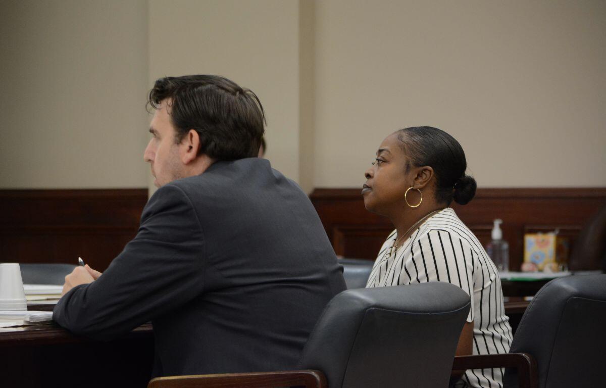 Two adults sit at a dark wood table in a formal setting; a man in a dark suit faces left and a woman with a striped blouse and hoop earrings sits beside him, both looking ahead.