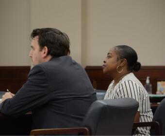 Two adults sit at a dark wood table in a formal setting; a man in a dark suit faces left and a woman with a striped blouse and hoop earrings sits beside him, both looking ahead.