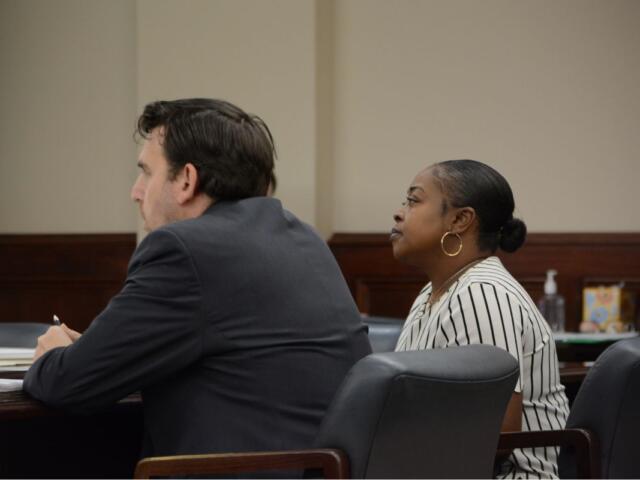 Two adults sit at a dark wood table in a formal setting; a man in a dark suit faces left and a woman with a striped blouse and hoop earrings sits beside him, both looking ahead.