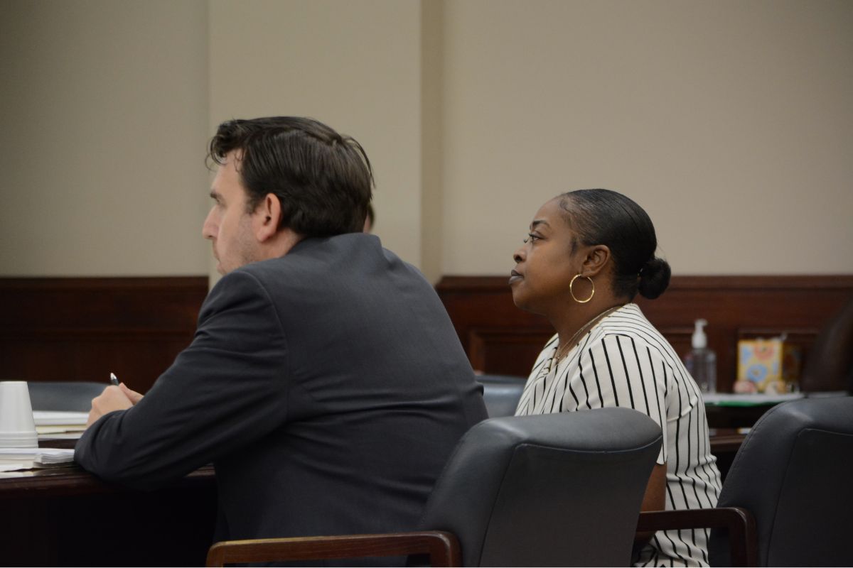 Two adults sit at a dark wood table in a formal setting; a man in a dark suit faces left and a woman with a striped blouse and hoop earrings sits beside him, both looking ahead.