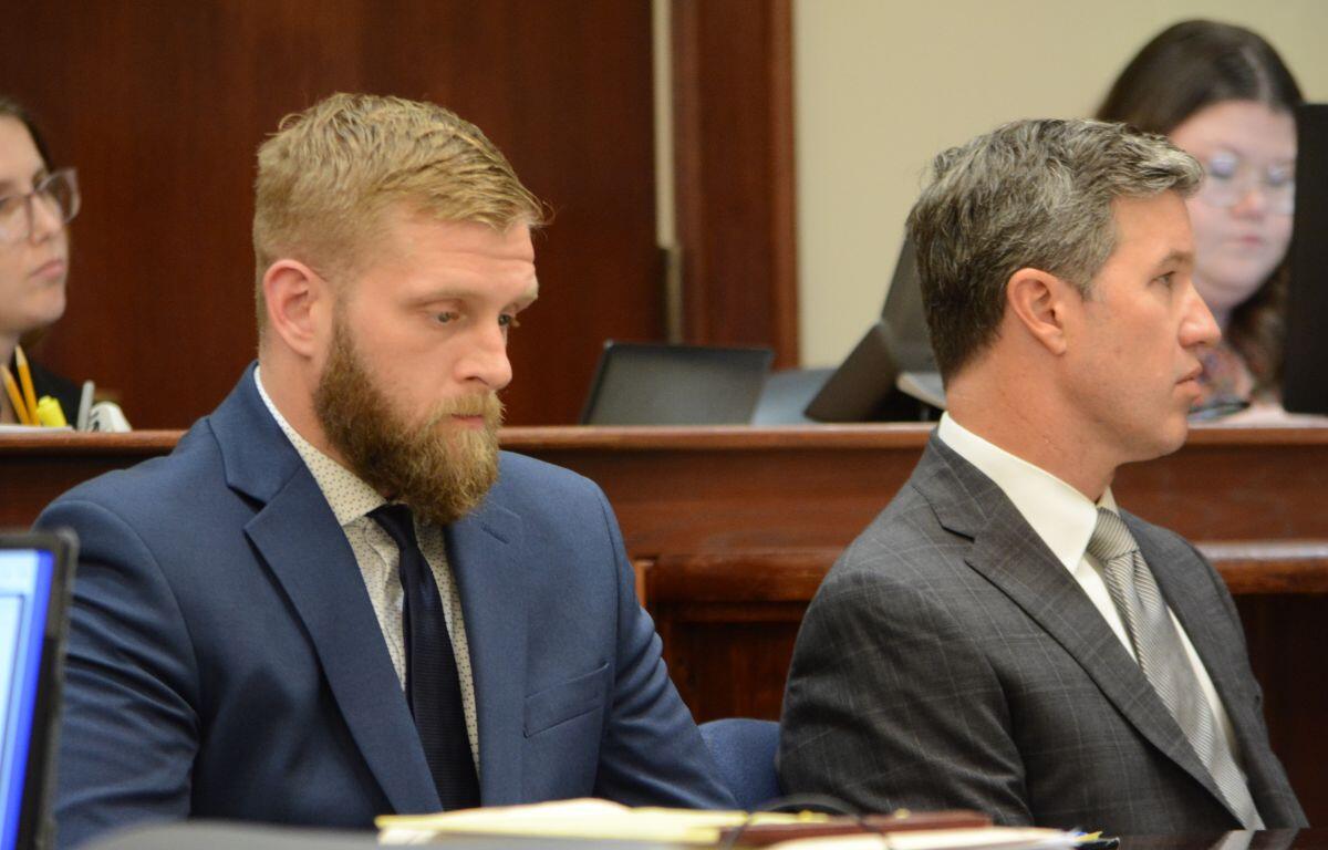 Two men in suits sit side by side at a formal bench, one with a beard in a blue suit and the other in a gray check suit.