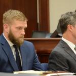 Two men in suits sit side by side at a formal bench, one with a beard in a blue suit and the other in a gray check suit.