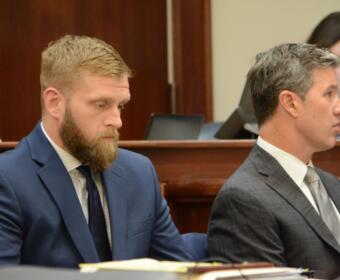 Two men in suits sit side by side at a formal bench, one with a beard in a blue suit and the other in a gray check suit.