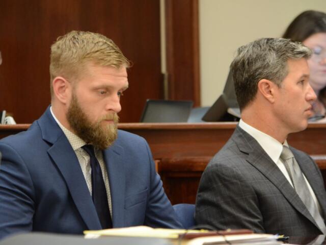 Two men in suits sit side by side at a formal bench, one with a beard in a blue suit and the other in a gray check suit.
