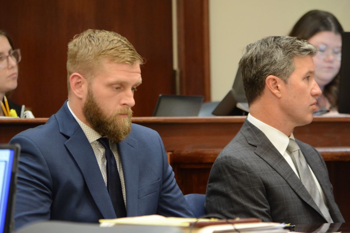 Two men in suits sit side by side at a formal bench, one with a beard in a blue suit and the other in a gray check suit.