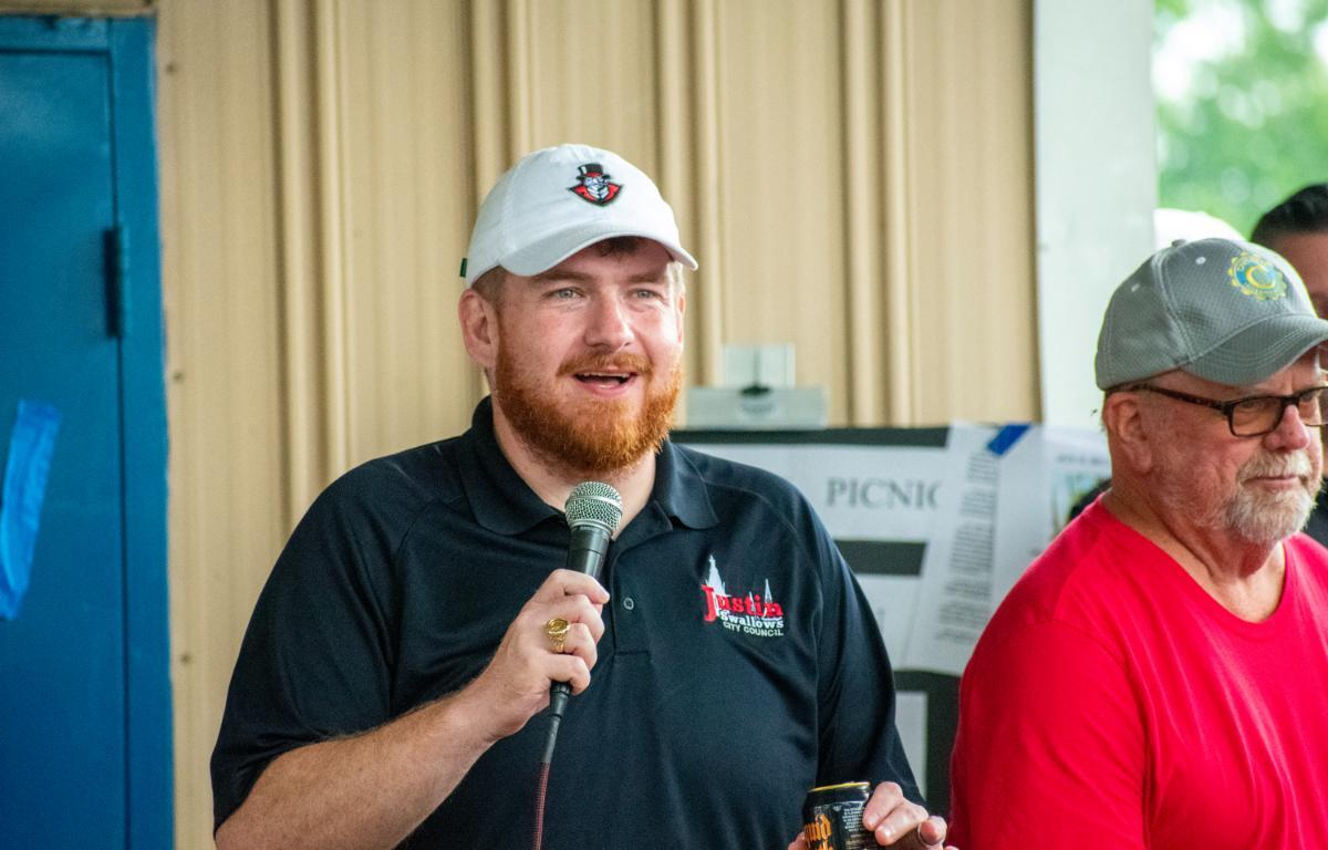 Bearded man in a white cap and black polo speaks into a microphone at an outdoor event, holding a can in his other hand.