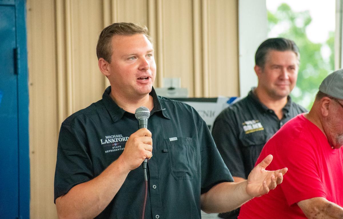 Man in a dark short-sleeve shirt speaks into a handheld microphone at an indoor event, with two men in the background.