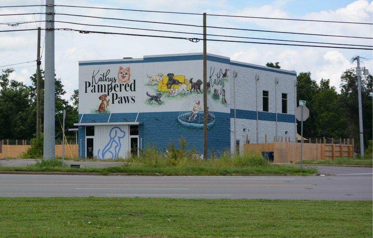 Two-story blue-gray building with a colorful dog mural that reads 'Kathy's Pampered Paws' and shows dogs at play and a fountain scene.