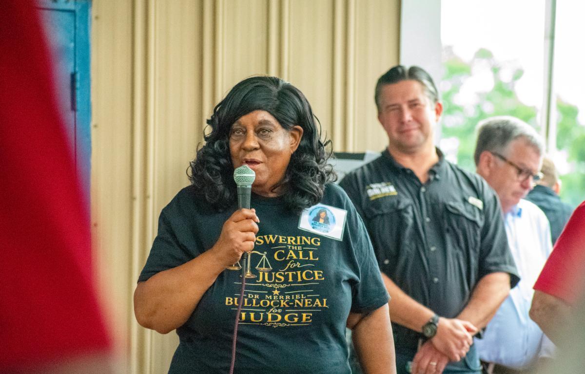 A Black woman speaks into a wired microphone at a justice-themed event, wearing a dark T-shirt with 'Answering the Call for Justice' and a name badge.